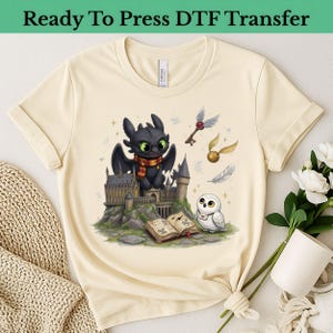 May include: A cream-colored t-shirt featuring a graphic print of a black dragon wearing a red and yellow scarf, a white owl, a castle, an open book, a golden snitch, and a winged key. The design includes small stars and sparkles.