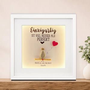 Könnte beinhalten: Ein quadratischer weißer Bilderrahmen mit einem Steinbild, das eine Figur auf einem Holzsteg zeigt, die einen roten herzförmigen Luftballon hält. Der Text lautet: Einzigartig ist viel besser als perfekt, Bleib so, wie du bist! Steffi, in Dunkelbraun.