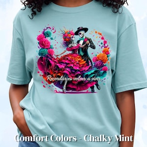 Peut inclure: Un t-shirt vert menthe clair avec un motif de deux squelettes dansant, entourés de roses vibrantes roses, orange et bleu sarcelle. Le texte "Recordar es volver a vivir" est imprimé en écriture blanche sous les personnages, avec "Comfort Colors - Chalky Mint" en dessous.
