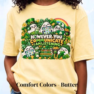 May include: A butter yellow t-shirt featuring a graphic of four ghosts surrounded by shamrocks, gold coins, and a rainbow. The text reads: HOWEVER YOU COMMUNICATE I AM LISTENING, with books labeled AAC, PECS, SPEECH, SIGN LANGUAGE, and SOCIAL STORIES.