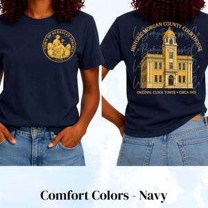 May include: A navy blue t-shirt featuring a gold circular graphic on the front chest that reads Town of Berkeley Springs 1776. The back displays a gold illustration of the Historic Morgan County Courthouse with the text Original Clock Tower Circa 1907.