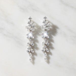 May include: A pair of silver-coloured drop earrings with a floral design. Each earring features a series of sparkling, clear crystals arranged in a cascading pattern. The earrings are designed to dangle and catch the light, creating a glamorous effect.