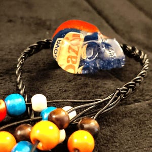 May include: A unique necklace featuring a blue skull pendant with red and white accents. The skull is made from a repurposed material with the word "Goya" visible. The necklace includes a black braided cord and colorful wooden beads.