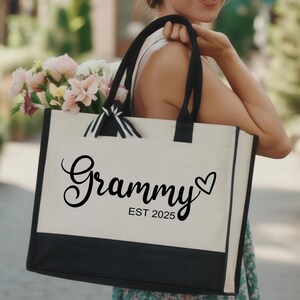May include: Cream-colored tote bag with black accents and handles. The bag features the word "Grammy" in a script font, a heart symbol, and the text "EST 2025". The bag is filled with pink and white flowers.