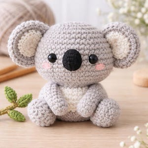 Könnte beinhalten: Ein handgehäkelter Koala-Bär in Grau-, Weiß- und Schwarztönen. Der Koala hat große, runde Ohren mit cremefarbenen Zentren, schwarze Augen und rosafarbene Wangen. Er sitzt auf einer hellen Holzoberfläche.