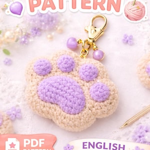 Könnte beinhalten: Ein gehäkelter Pfotenabdruck-Schlüsselanhänger in Beige und Lavendel. Der Pfotenabdruck ist an einem goldfarbenen Verschluss mit Lavendelperlen befestigt. Das Bild enthält auch den Text "CROCHET PATTERN", "PDF PATTERN" und "ENGLISH TUTORIAL".