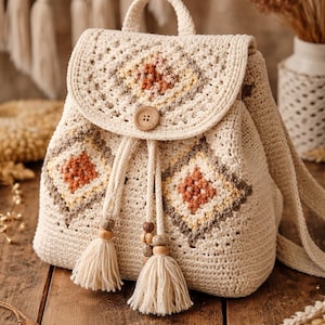 Könnte beinhalten: Cremefarbener Häkelrucksack mit Klappenverschluss und Holzknopf. Die Tasche hat geometrische Rautenmuster in Braun- und Beigetönen. Quasten und Holzperlen hängen am Kordelzugverschluss. Oben befindet sich ein Griff.