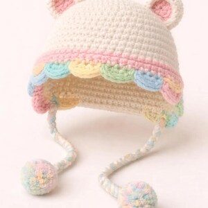 Könnte beinhalten: Gehäkelte Baby-Mütze in Creme mit rosa gefütterten Ohren. Die Mütze hat einen gewellten Besatz in Pastellfarben: Rosa, Gelb, Grün und Blau. Passende Pastell-Pompons hängen an den Bändern der Mütze.