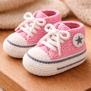 May include: Hand-crocheted baby sneakers in pink and white. The shoes feature white laces, a grey stripe around the sole, and a star patch. They are displayed on a wooden surface. A charming gift idea.