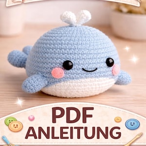 Puede incluir: Una ballena azul y blanca de ganchillo con una cara sonriente, mejillas rosadas y pequeñas aletas. La imagen incluye el texto "HÄKELANLEITUNG" y "PDF ANLEITUNG". También se ven botones decorativos e hilo.