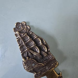 Peut inclure: Un heurtoir de porte en laiton présentant un relief détaillé d'un voilier avec plusieurs mâts et voiles. Le métal présente une patine sombre et vieillie avec des reflets sur les surfaces en relief du navire et les bords festonnés décoratifs.