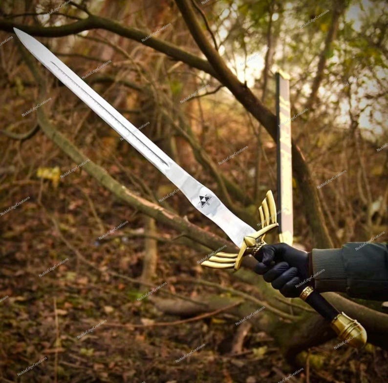 May include: A silver-colored sword with a gold-colored hilt and a black handle. The blade is long and straight, with a triangular emblem near the hilt. The sword is held against a backdrop of trees.