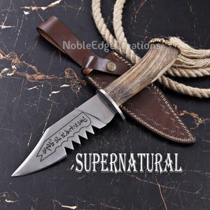 May include: A fixed-blade knife with a textured antler handle and a silver-toned steel blade featuring a serrated edge and engraved runic symbols. The knife is accompanied by a brown leather sheath with a metal snap button closure.
