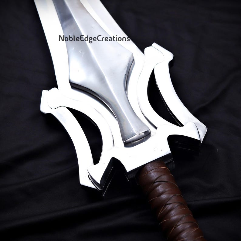 May include: A close-up of a silver sword with a brown leather-wrapped handle. The sword features a wide, double-edged blade and a distinctive guard design. The text "NobleEdgeCreations" is visible on the blade.