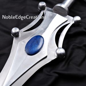 May include: Close-up of a silver sword with a blue oval gemstone set in the center of the hilt. The sword has a polished finish and a decorative design with rounded accents. The text "NobleEdgeCreations" is visible.