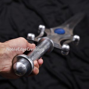 May include: A silver-colored sword with a blue gem in the center of the blade. The handle is cylindrical and held in a hand. The sword has a crossguard with rounded protrusions. The text "NobleEdgeCreations" is visible.