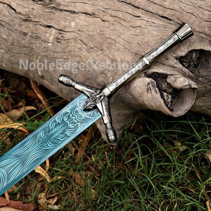 Könnte beinhalten: Ein Schwert mit einem metallisch silbernen Griff, der mit komplizierten Gravuren und einem Knauf versehen ist. Die Klinge ist hellblau und weist ein dunkles, wirbelndes Muster auf. Der Text NobleEdgeCreations ist auf der Klinge nahe dem Griff zu sehen.