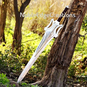 May include: A large, silver sword with a decorative hilt and a brown leather-wrapped handle. The blade is long and pointed, with a polished finish. The sword is leaning against a tree trunk. The text "Noble Edge Creations" is visible.