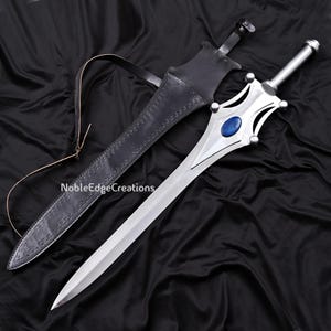 May include: A silver sword with a blue gem in the center of the hilt, resting next to a black leather sheath. The sword has a decorative handle and a long, sharp blade. The sheath has a strap and is embossed with a pattern. The text "NobleEdgeCreations" is visible.