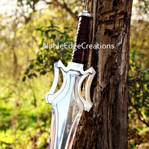 May include: A silver sword with a brown leather-wrapped handle rests against a tree. The sword features a unique hilt design and a polished blade. The text "NobleEdgeCreations" is visible on the tree.