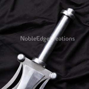 May include: Close-up of a silver sword with a cylindrical handle and a decorative crossguard. The sword has a polished finish and the text "NobleEdge Creations" is visible.