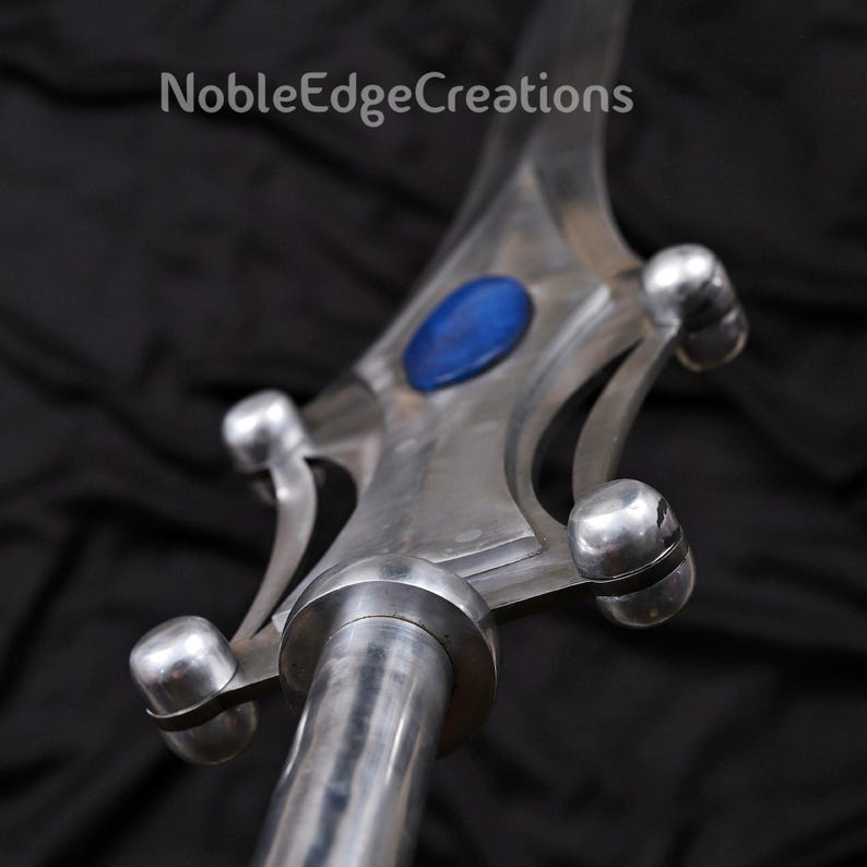 May include: A close-up of a silver metal sword with a blue oval-shaped gem in the center of the hilt. The sword has a polished finish and rounded accents. The text "NobleEdgeCreations" is in the upper left corner.