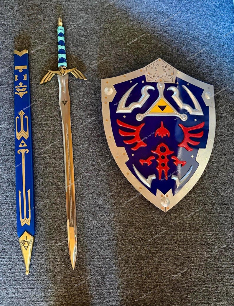 May include: A prop sword, shield, and scabbard set. The sword has a gold hilt and blade, with a blue and teal handle. The shield is blue with silver trim and red and gold accents. The scabbard is blue and gold.