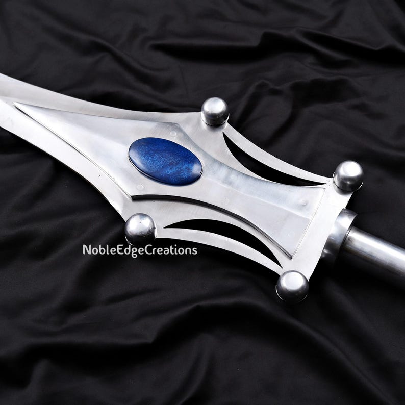 May include: Close-up of a silver sword with a blue oval stone in the center of the blade. The sword has a decorative design with rounded silver accents. The text "NobleEdgeCreations" is visible on the blade.