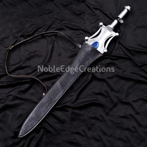 May include: A silver sword with a blue gem in the hilt, resting in a black leather sheath. The sword has a decorative handle and a leather strap. The text "NobleEdgeCreations" is visible on the background.