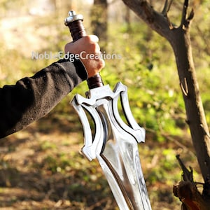 May include: A polished silver sword with a decorative hilt and a brown leather-wrapped handle is held in a person's hand. The sword's blade reflects the surroundings. The text "Noble Edge Creations" is visible on the image.