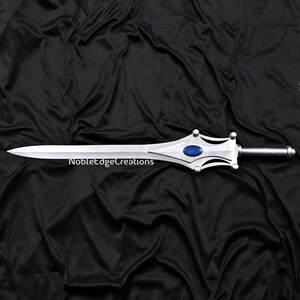 May include: A silver sword with a pointed blade and a decorative hilt featuring a blue gem. The text "NobleEdgeCreations" is printed on the blade. The sword is displayed against a black background.