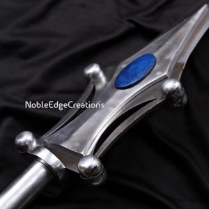 May include: Close-up of a silver spearhead with a blue oval inlay. The spearhead has a polished finish and a decorative design. The text "NobleEdgeCreations" is visible on the side. The spear is set against a dark background.