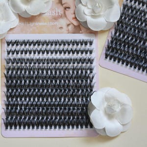 May include: A rectangular tray containing multiple rows of individual black false eyelash clusters arranged in a grid pattern. The tray features a printed background with a face and cat illustration, and white fabric flowers are placed around the tray.