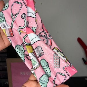May include: Pink headband with a repeating pattern of medical equipment, including stethoscopes, syringes, pills, and Crocs. The headband is made of a soft fabric and is designed to be worn on the head. The text "RN GIRL" is visible on a pink box in the background.