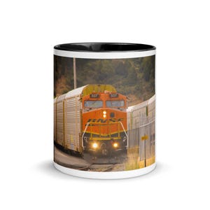 May include: White ceramic mug with a black interior, featuring an image of an orange BNSF train engine with the number 507. The train is traveling on a track with a backdrop of green trees and a cloudy sky.