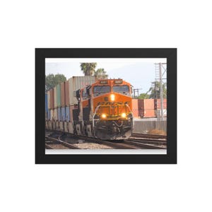 Op de afbeelding: Een oranje BNSF-goederentreinlocomotief met nummer 3257 die een reeks veelkleurige vrachtcontainers over metalen rails trekt. De trein is voorzien van heldere koplampen en een zwarte lijst, gepresenteerd als ingelijste wanddecoratie.