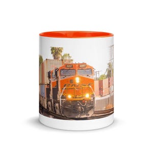 May include: A white ceramic mug with an orange interior rim featuring a photographic print of an orange BNSF freight train locomotive numbered 3257, pulling several shipping containers along tracks with palm trees visible in the background.
