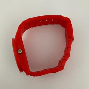 May include: A bright red wristwatch with a rectangular face and a matching segmented band. The watch has a simple design with a silver-colored button on the side. The band is made of connected rectangular links.