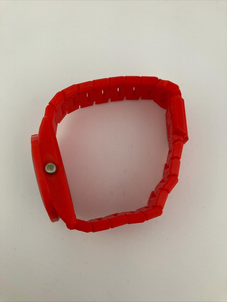 May include: A bright red wristwatch with a rectangular face and a matching segmented band. The watch has a simple design with a silver-colored button on the side. The band is made of connected rectangular links.
