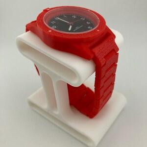 May include: A red wristwatch with a black watch face is displayed on a white stand. The watch has a textured red band and casing. The stand has a curved design.
