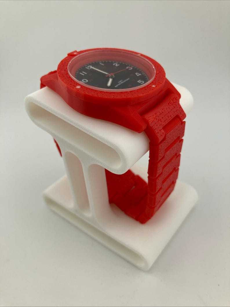 May include: A red wristwatch with a black watch face is displayed on a white stand. The watch has a textured red band and casing. The stand has a curved design.