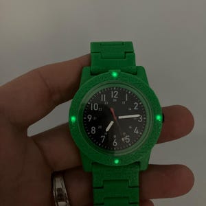 May include: A green wristwatch with a black watch face. The watch has a green band and a textured bezel. The watch face displays the numbers 1 through 12, with a red second hand. The watch is held by a hand.