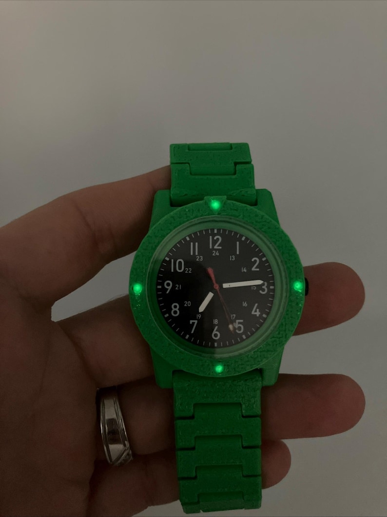 May include: A green wristwatch with a black watch face. The watch has a green band and a textured bezel. The watch face displays the numbers 1 through 12, with a red second hand. The watch is held by a hand.