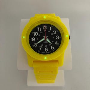 May include: A bright yellow wristwatch with a black watch face. The watch has white numerals and hands, with a red second hand. The watch band is also yellow. The watch is displayed on a white stand.