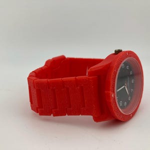 May include: A red wristwatch with a black watch face. The watch has a square case and a matching red band. The watch face displays the time with white numerals and hands. The band is made of rectangular links.