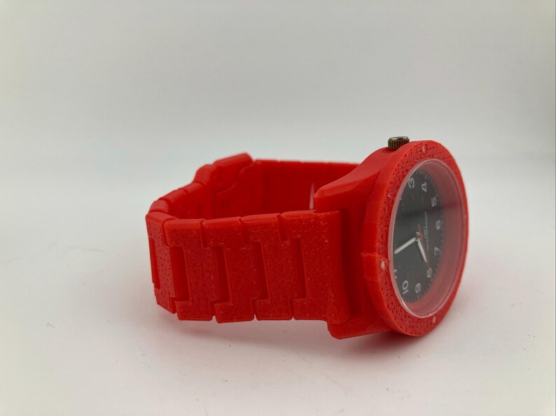 May include: A red wristwatch with a black watch face. The watch has a square case and a matching red band. The watch face displays the time with white numerals and hands. The band is made of rectangular links.