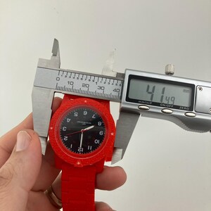 May include: A red wristwatch with a black face is measured with a digital caliper. The watch face displays white numerals and hands. The caliper reads 41.49 mm, approximately 1.63 inches.