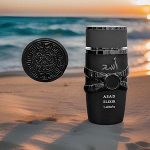 May include: Black perfume bottle with silver accents and a matching round cap. The bottle has the word "ASAD ELIXIR Lattafa" printed on it. The background shows a beach scene with the ocean and sunset.