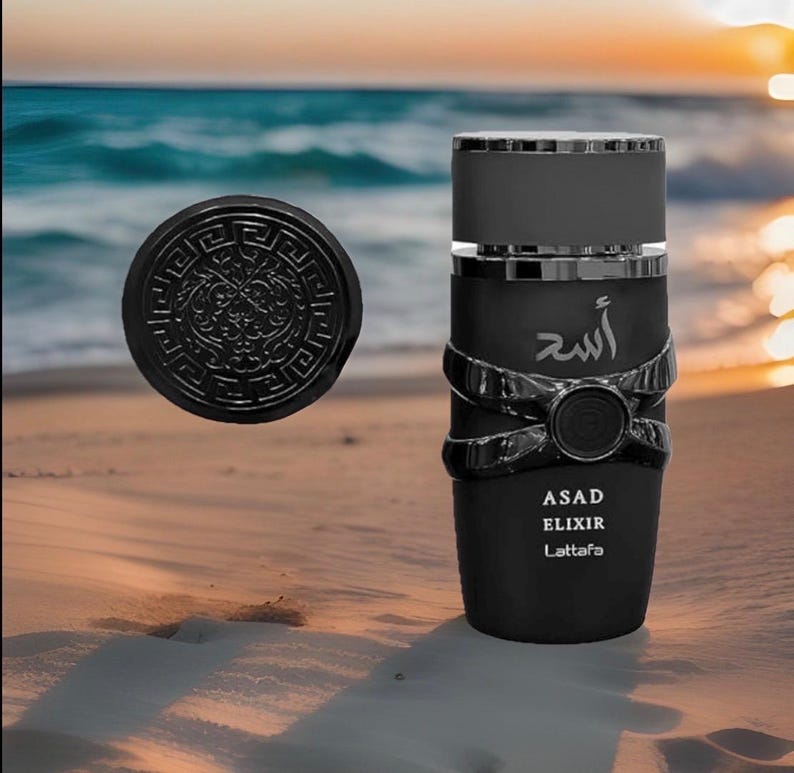 May include: Black perfume bottle with silver accents and a matching round cap. The bottle has the word "ASAD ELIXIR Lattafa" printed on it. The background shows a beach scene with the ocean and sunset.