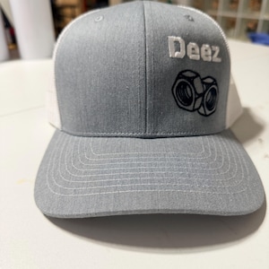 May include: Gray trucker hat with a white mesh back and a curved brim. The front of the hat features the word "Deez" in white block letters above a black and white embroidered image of two nuts.